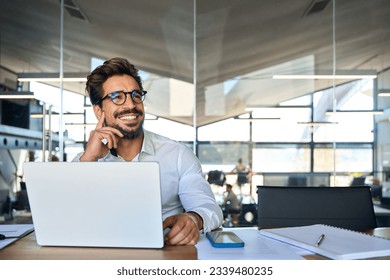 Happy Latin business man working at laptop in office looking away. Happy young businessman professional using computer sitting at desk thinking of corporate technology ai solutions. Copy space - Powered by Shutterstock