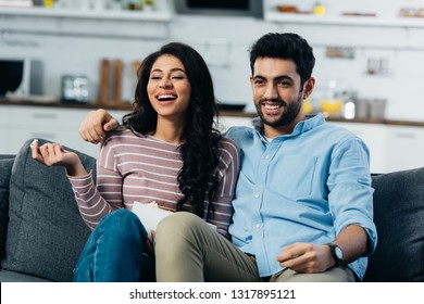 Happy Latin Couple Watching Tv At Home With Bowl Of Popcorn