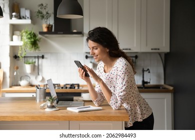 Happy Latin freelance employee working from home, using online app on smartphone at laptop in kitchen, smiling at video call, texting on social media chats, reading article. Digital life concept - Powered by Shutterstock