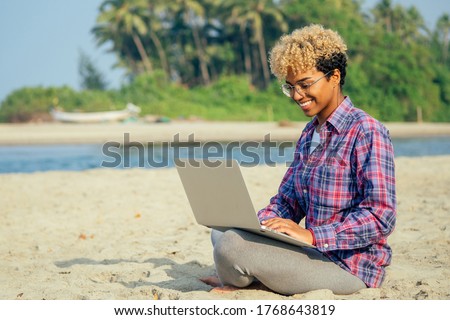 happy latin freelancer woman with blonde afro curls sitting at empty beach with laptop palm trees background. remote dream best work concept