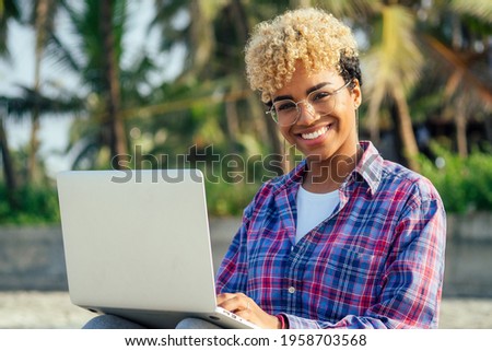 happy latin freelancer woman with blonde afro curls sitting at empty beach with laptop palm trees background. remote dream best work concept