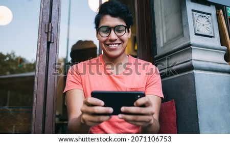 Happy Latin hipster guy in stylish spectacles for provide eyes correction enjoying leisure time for playing online games via palmtop technology, cheerful male user connecting to 4g on smartphone