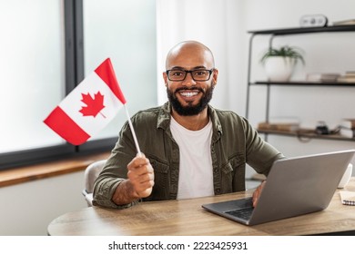 Happy latin man sitting with flag of Canada and using laptop computer, sitting at desk at home. Modern online foreign education, emigration and citizenship, working abroad concept - Shutterstock ID 2223425931