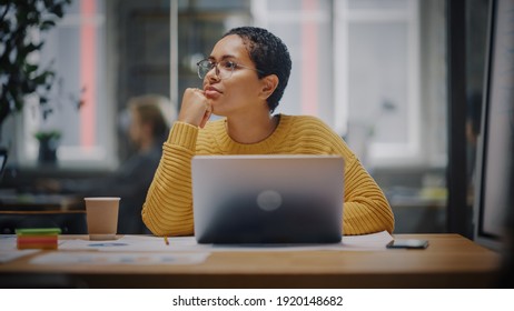 Happy Latin Marketing Specialist In Glasses Working On Laptop Computer In Busy Creative Office. Beautiful Diverse Female Project Manager Hesitating And Thinking Before Answering An Email To Colleague.