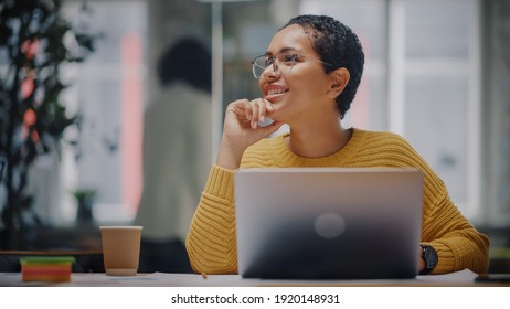 Happy Latin Marketing Specialist In Glasses Working On Laptop Computer In Busy Creative Office. Beautiful Diverse Female Project Manager Hesitating And Thinking Before Answering An Email To Colleague.