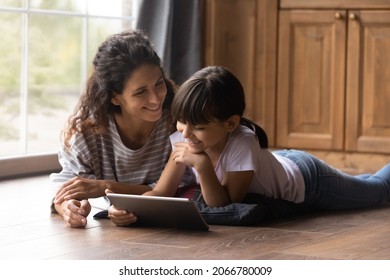 Happy Latin mom and girl resting on heating parquet floor in kitchen, using tablet, laughing, watching movie, making video call, reading ebook on internet together. Mother and daughter at home - Powered by Shutterstock