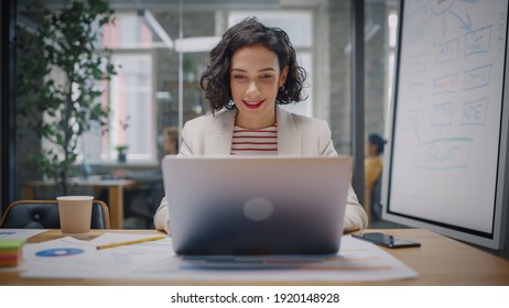 Happy Latin Project Manager with Dark Wavy Hair Working on Laptop in Busy Creative Office. Beautiful Diverse Female Ad Manager Comparing Reports and Graphs on Paper and on Her Computer. - Powered by Shutterstock