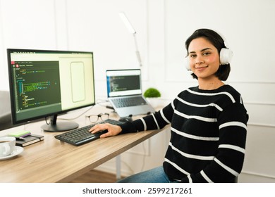 Happy latin software developer working remotely and coding a mobile application while wearing headphones and sitting at her desk