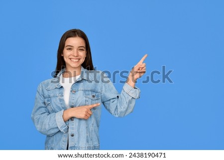Happy Latin teen girl pointing aside standing on blue background. Smiling pretty freckled gen z brunette Hispanic teen student wearing denim outfit looking at camera presenting product or service ad.
