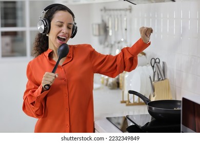 Happy latin woman cooking, smelling and tasting food while dancing in kitchen. Beautiful young female with headphone preparing delicious meal while listen music at home. Healthy and lifestyle concept - Shutterstock ID 2232349849
