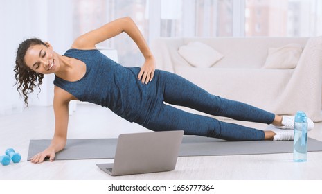 Happy Latin Woman Doing Side Plank Exercise While Watching Workout Video On Laptop At Home