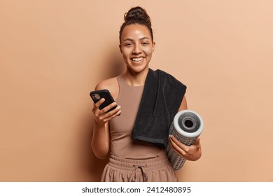 Happy Latin woman dressed in activewear uses foam roller for doing exercises to relieve back pain holds smartphone being always in touch smiles happily isolated on brown background. Fascia stretching - Powered by Shutterstock