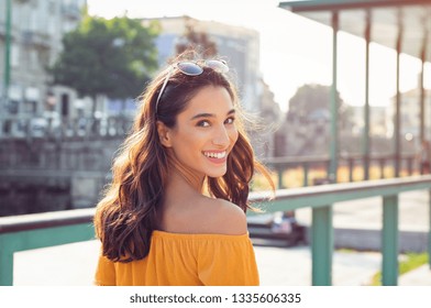 Happy latin woman looking behind while walking on street. Beautiful young woman enjoying vacation during summer. Cheerful girl in smart casual walking and looking at camera while smiling.