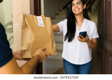 Happy latin woman receiving a food delivery order in a brown paper bag at the doorway of her home