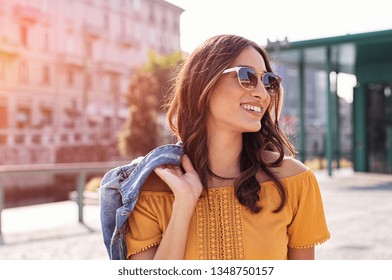 Happy latin woman walking on the street on a bright sunny day. Cheerful stylish girl with sunglasses enjoying the spring. Young woman wearing shades while looking away in the afternoon.