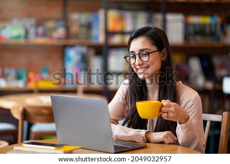 Happy latin woman working online at cafeteria, using laptop computer and drinking coffee, copy space. Excited arab female freelancer typing on laptop, holding mug of hot beverage