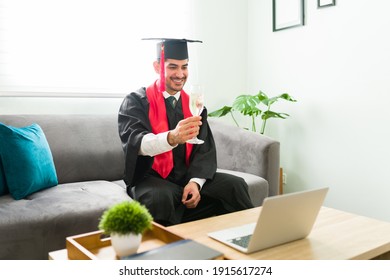 Happy Latin Young Man Making A Toast With A Glass Of Wine During His Virtual Graduation Ceremony. Male Graduate Celebrating His Academic Success In A Video Call Because Of The Coronavirus Pandemic