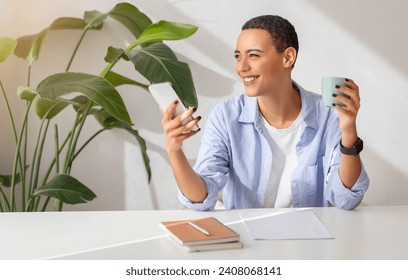 Happy latin young woman in a light blue shirt multitasking with a smartphone in one hand and a mug in the other at a modern workspace with a notepad on the desk, enjoy chat - Powered by Shutterstock