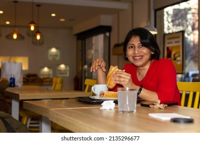 Happy Latina Woman Having Breakfast In A Cafe Having A Sandwich And Coffee.