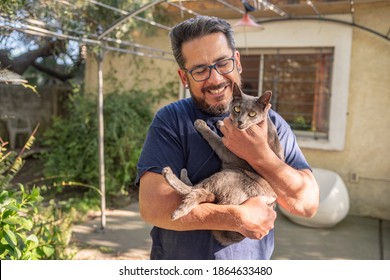 A Happy Latino Man Living With HIV Holding A Gray Cat Outside His Home