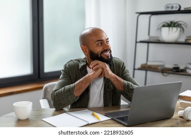 Happy Latino Man Sitting At Table In Front Of Laptop Computer At Home, Looking Aside And Thinking. Positive Freelancer Man Daydreaming While Working Online, Using Portable Pc Indoors