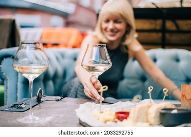 Happy and laughing adult mature woman sitting in bar outdoors with wine glasses and blurry restaurant background scene, drinking white wine and eating cheese. Summer sunny day on patio.