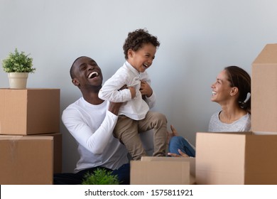 Happy laughing african american young man rising up joyful mixed race cute little son, having fun with wife. Overjoyed smiling multiracial family playing near carton packages, moving in first home.