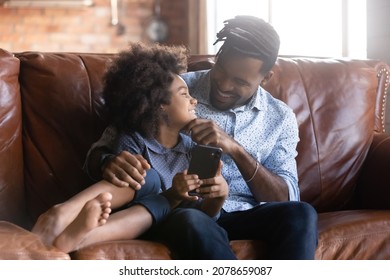 Happy laughing African ethnicity cute child girl using funny smartphone applications with caring young biracial father, spending time online playing mobile games or shopping, entertaining at home.
