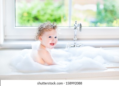 Happy Laughing Baby Girl Having Fun Playing With Water And Foam In A Kitchen Sink Next To A Window