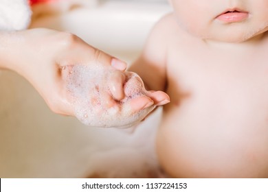 Happy Laughing Baby Taking A Bath Playing With Foam Bubbles. Little Child In A Bathtub. Infant Washing And Bathing. Unrecognizable Mother Bathing Her Baby In White Small Plastic Bat