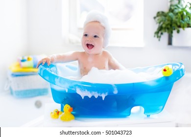 Happy Laughing Baby Taking A Bath Playing With Foam Bubbles. Little Child In A Bathtub. Smiling Kid In Bathroom With Colorful Toy Duck. Infant Washing And Bathing. Hygiene And Care For Young Children.