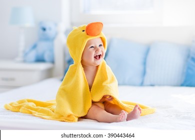 Happy laughing baby wearing yellow hooded duck towel sitting on parents bed after bath or shower. Clean dry child in bedroom. Bathing and washing of little kids. Children hygiene. Textile for infants. - Shutterstock ID 390443464