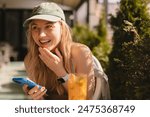 Happy laughing blonde woman in cap sitting in cafe and drink cocktail. Girl hold mobile phone read some funny news, watching memes or reading jokes and laughing. Have a good time.