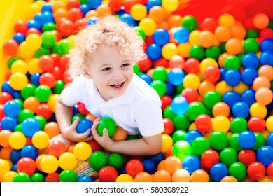 Happy laughing boy having fun in ball pit on birthday party in kids amusement park and indoor play center. Child playing with colorful balls in playground ball pool. Activity toys for little kid