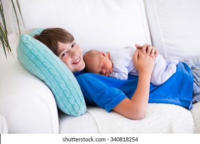 Happy Laughing Boy Holding His Sleeping Newborn Baby Brother. Siblings With Big Age Difference. Children Playing At Home On A White Couch. New Born Child Taking A Nap. Kids Bonding. Family Love. 