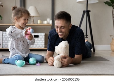 Happy Laughing Caring Dad And Daughter Kid Playing Together On Heating Floor, Relaxing In Living Room, Having Fun At Tea Party With Teddy Bear, Toy Cups, Pot, Dish. Fatherhood, Family Playtime