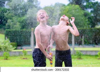 Happy Laughing Children, Two Young School Boys, Enjoying Hot Sunny Summer Vacation Day Playing Outdoors In Garden At The Backyard Of The House Running On Green Lawn And Spraying Water From The Hose