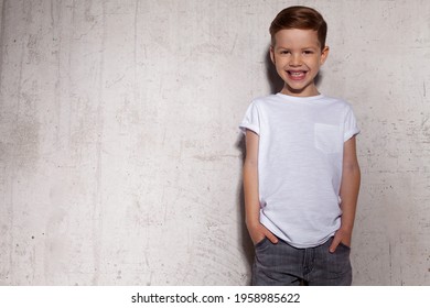 Happy Laughing Cool Little Boy In White T-shirt Posing On Background Of Grunge Concrete Wall With Copy Space For Ad. Smiling Stylish Fashionable Schoolboy Near Gray Wall. Children's Style And Fashion.