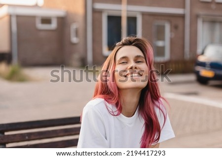 Happy laughing girl with pink hair sitting on bench and look positive. Outdoor good mood woman in city. 