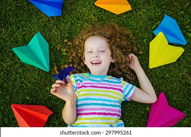 Happy Laughing Girl Throwing Paper Airplane In Green Grass At Summer Park. Happy Childhood, Travel, Vacation Concept. Top View.