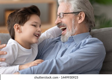 Happy Laughing Grandfather Wearing Glasses Playing, Hugging Adorable Little Grandson, Excited Granddad And Preschool Grandchild Boy Having Fun At Home, Sitting On Comfortable Couch, Close Up