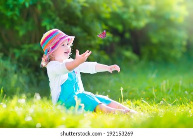 Happy laughing little girl wearing a blue dress and colorful straw hat playing with a flying butterfly having fun in the garden on a sunny summer day