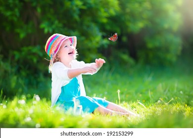 Happy Laughing Little Girl Wearing A Blue Dress And Colorful Straw Hat Playing With A Flying Butterfly Having Fun In The Garden On A Sunny Summer Day