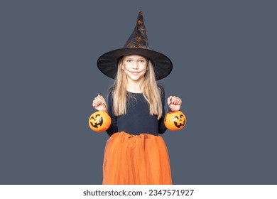 A happy laughing little girl in a witch costume and makeup holds pumpkin baskets for Halloween treats on a dark background. Children's Halloween