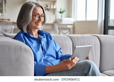 Happy laughing mid aged senior woman sitting on couch with digital tablet gadget on video call with family doctor. Older female reading ebook or attenting video meeting online.
