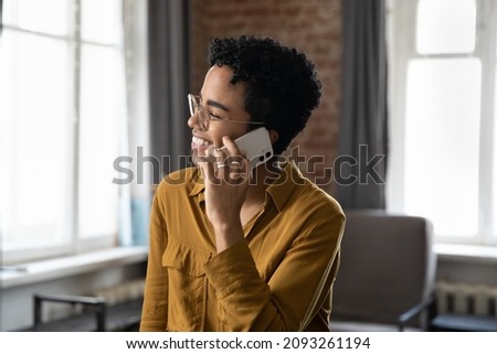 Happy laughing millennial African American woman in eyewear holding pleasant distant phone call conversation, sharing good life news, discussing issues, communicating distantly in modern home office.