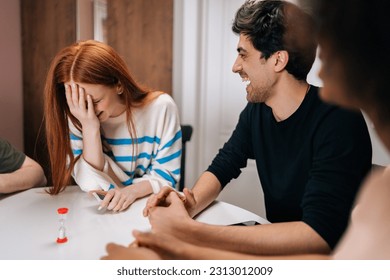 Happy laughing multiethnic friends having play cards games for time, enjoying board games activity for entertainment at home. Cheerful diverse men and women playing UNO board game sitting at table.