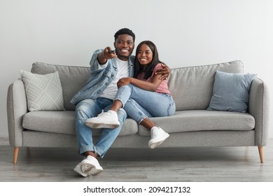 Happy laughing relaxed millennial African American woman and man watching tv in living room interior, holding remote control and hugging. Smiling couple enjoying film and movie evening together - Powered by Shutterstock