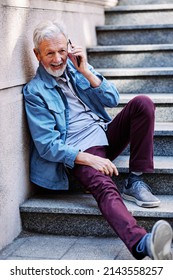 A Happy Laughing Senior Man Is Sitting On The Stairs Outdoors And Flirting On The Phone.