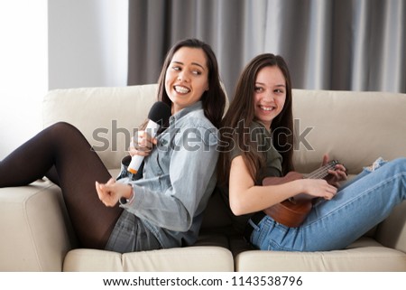 Happy laughing sisters on the couch in the living room. One is playing at the ukulele and the other is singing at the microphone